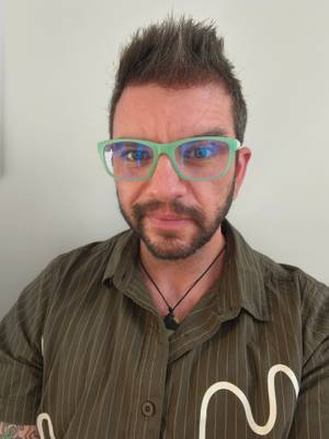 A white middle-aged man with dark hair and beard looks at the camera. He's wearing a green shirt and green glasses.