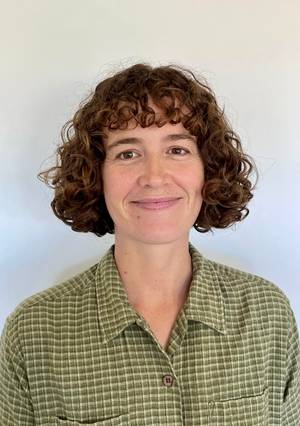 Ella smiles at the camera against a white background. She has short curly hair and they are wearing a check shirt
