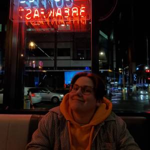 A Pākēha person with short hair and glasses smiles at the camera. They're outside, wearing a hoodie and jacket.