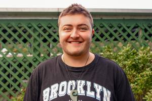 A man with short dark hair smiles at the camera. He's wearing a pounamu and a black t-shirt