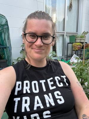 A Pākēha woman with long blond hair and glasses smiles at the camera. She is outside in a garden. Her shirt reads "Protect trans kids"