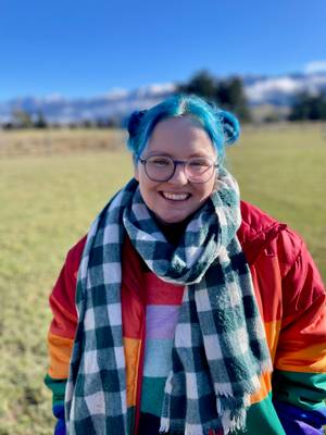 A person with blue hair and glasses smiles at the camera. They're outside, with a green and white checked scarf and orange jacket