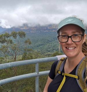 A woman wearing glasses and a visor smiles at the camera. She is outdoors with a background of forest and cliffs in the distance