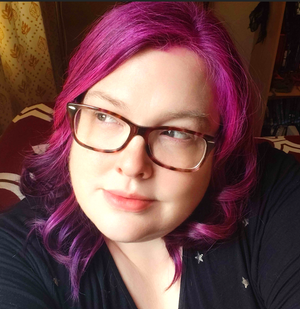 A woman with pink/purple hair and glasses looks away from the camera, lit from sunlight outside